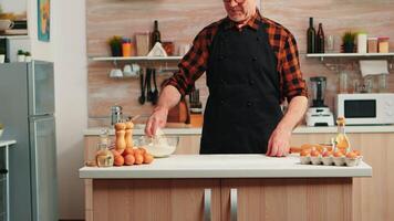 Elderly baker flouring table to bake and make delicious cookies. Retired senior chef with bonete and apron in uniform sprinkling sieving sifting raw ingredients by hand baking homemade pizza, bread video