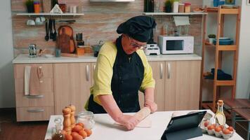 Senior Frau mit Bonete suchen Rezept beim das Digital Laptop. im Ruhestand Dame folgenden kulinarisch Rat auf Tablette, Lernen Kochen Lernprogramm auf Sozial Medien, mit hölzern rollen Stift Bildung das Teig. video