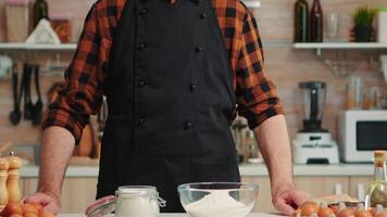 Senior padeiro sorridente olhando às Câmera dentro cozinha com Comida ingredientes em mesa para cozimento. aposentado idosos chefe de cozinha com bonete pronto para cozinhar caseiro pão, bolos e massa com trigo farinha e ovos video