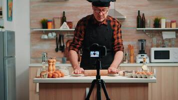 Baker using wooden rolling pin for dough in front camera recording new cooking episode. Old blogger chef influencer using internet technology communicating on social media with digital equipment video