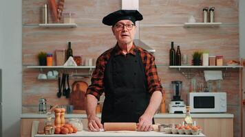 Elderly man rolling dough at home infront of camera explaining recipe step by step. Retired blogger chef influencer using internet technology communicating on social media with digital equipment video