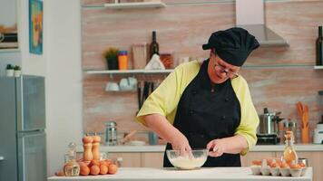 mature femme boulanger mélange par main fissuré des œufs avec farine dans Accueil cuisine Suivant traditionnel recette. retraité personnes âgées chef avec bonte pétrissage dans verre bol Pâtisserie Ingrédients cuisson fait maison gâteau video