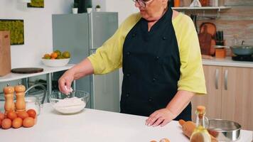 Frau Koch nehmen Weizen Mehl von Glas Schüssel und Sieben auf Tisch. im Ruhestand Senior Bäcker mit Bonete und Uniform streuen, Sichtung, Verbreitung rew Zutaten Backen hausgemacht Pizza und Brot. video