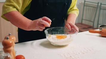 Close up of hands woman adding eggs in flour for baking. Elderly pastry chef cracking egg on glass bowl for cake recipe in kitchen, mixing by hand, kneading ingredients prreparing homemade cake video