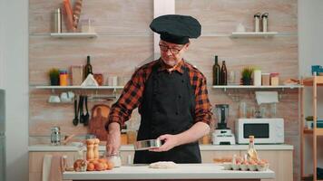 Baker with bonete and apron using metallic sieve preparing bakery products at home. Happy elderly chef with kitchen uniform mixing, sprinkling, sieving raw ingredients to baking traditional bread video