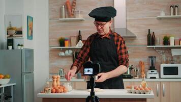 Cheerful elderly baker man filming cooking vlog in home kitchen. Retired blogger chef influencer using internet technology communicating, shooting blogging on social media with digital equipment video