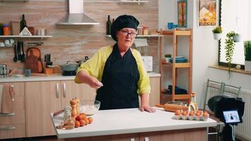 Elderly social media influencer cooking bread using camera on tripod sitting in the kitchen. Retired blogger chef using internet technology communicating, shooting blogging with digital equipment video