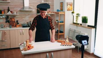 Old man shooting culinary using camera on tripod in the kitchen. Retired blogger chef influencer using internet technology communicating, shooting blogging on social media with digital equipment video