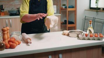 Old lady chef making the shape for pizza at home in kitchen. Skillful retired elderly baker wearing uniform forming the dough on a floured surface and kneading it spinning, tossing throwing it up video