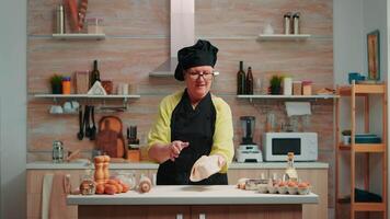 Woman with bonete having fun cooking at home in modern kitchen, making dust with flour. Skillful retired elderly chef wearing uniform spinning and tossing pizza dough throwing it up video