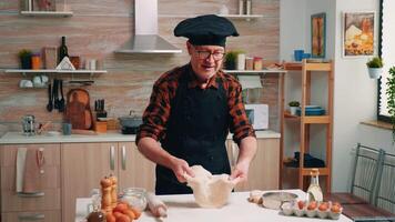 Cheerful retired chef in uniform spinning and tossing pizza dough throwing it up, forming on a floured surface and kneading it shaping. Skillful elderly baker wearing bonete cooking homemade cookies video