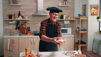 Happy elderly man throwing the pizza dough up. Skillful retired senior baker wearing uniform forming the dough on a floured surface and kneading it spinning and tossing up in home kitchen video