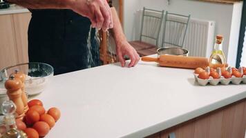 anziano uomo con bonete e cucina grembiule setacciare Farina su tavolo pronto per cucinando. pensionato anziano panettiere con uniforme aspersione, setacciare, diffusione rew ingredienti cottura al forno fatti in casa Pizza e pane. video