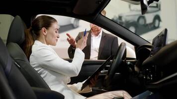 Car dealer giving keys to a young woman sitting inside the car. Joyful executive woman getting keys from new car. Happy auto owner buying transport at modern salon video