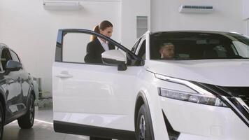 competente coche comerciante utilizando digital tableta para trabajo con clientela a sala de exposición. hembra hablando con gerente mientras comprando coche a concesión centro. hablando con gerente video