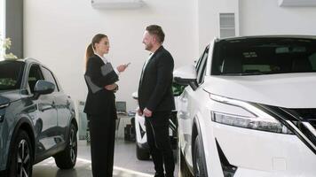 Competent car dealer using digital tablet for work with clients at showroom. Female talking with manager while buying car at dealership center. Talking with manager video