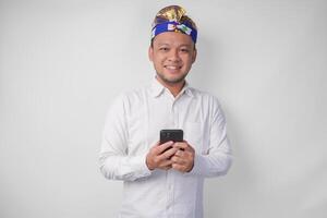 Excited Balinese man wearing white shirt and traditional headdress smiling happily while using smartphone, replying message, reading news photo