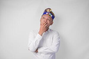 Confused Balinese man in traditional headdress called udeng thinking hard while touching chin with serious expression, isolated by white background photo