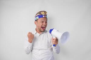 Overjoyed Balinese man in white shirt and traditional headdress shouting at megaphone feeling excited, isolated by white background photo