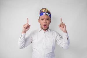 Surprised Balinese man wearing traditional headdress called udeng pointing up to the copy space upwards over isolated white background photo