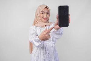 Portrait of young Asian Muslim woman wearing white dress and hijab smiling happily, pointing to the blank screen copy space on her phone and showing screen to the camera photo