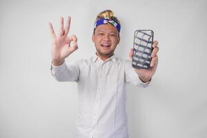 Happy young Balinese man wearing white shirt and traditional headdress presenting blank screen copy space on his phone while making okay, good, nice hand gesture photo