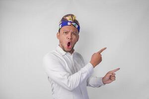 Surprised Balinese man in white shirt and traditional headdress called udeng pointing to the copy space on the left and right side photo