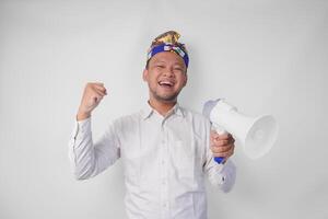 Energetic Balinese man in white shirt and traditional headdress shouting at megaphone while raising clenched fist, isolated by white background photo