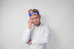 Confused Balinese man in traditional headdress called udeng thinking hard while touching chin with serious expression, isolated by white background photo