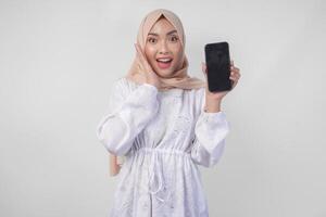 Portrait of young Asian Muslim woman wearing white dress and hijab smiling happily, pointing to the blank screen copy space on her phone and showing screen to the camera photo