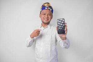 Smiling young Balinese man wearing white shirt and traditional headdress pointing to his smartphone, presenting blank screen copy space photo