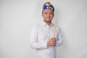 Balinese man wearing traditional headdress called udeng doing paying respect gesture while holding praying incense on isolated white background photo