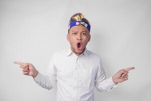 Surprised Balinese man in white shirt and traditional headdress called udeng pointing to the copy space on the left and right side photo