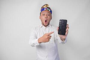 Surprised young Balinese man wearing white shirt and traditional headdress with shocked expression on face while pointing to his smartphone, presenting blank screen copy space photo