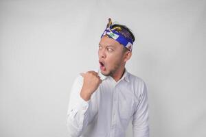 Surprised Balinese man in white shirt and traditional headdress called udeng pointing to the copy space on the left and right side photo