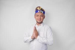 Smiling young Balinese man wearing traditional headdress called udeng doing greeting or welcome gesture, isolated over white background photo