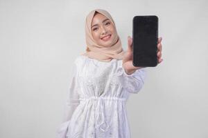 Portrait of young Asian Muslim woman wearing white dress and hijab smiling happily, pointing to the blank screen copy space on her phone and showing screen to the camera photo