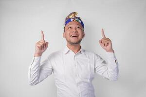 Surprised Balinese man wearing traditional headdress called udeng pointing up to the copy space upwards over isolated white background photo