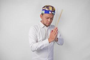 Balinese man wearing traditional headdress called udeng doing paying respect gesture while holding praying incense on isolated white background photo
