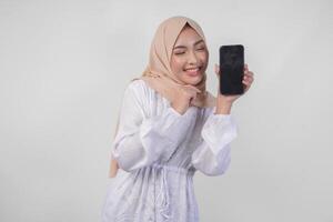 Portrait of young Asian Muslim woman wearing white dress and hijab smiling happily, pointing to the blank screen copy space on her phone and showing screen to the camera photo