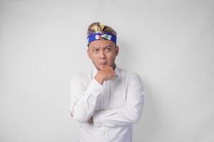 Confused Balinese man in traditional headdress called udeng thinking hard while touching chin with serious expression, isolated by white background photo