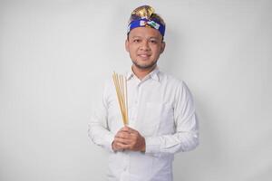 Balinese man wearing traditional headdress called udeng doing paying respect gesture while holding praying incense on isolated white background photo