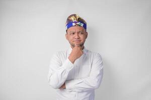 Confused Balinese man in traditional headdress called udeng thinking hard while touching chin with serious expression, isolated by white background photo