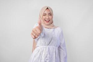 Attractive young Asian woman in white dress and hijab gesturing thumbs up for approval, good, excellent signs, isolated by white background photo