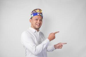 Attractive Balinese man in white shirt and traditional headdress called udeng pointing to the copy space on the left and right side while smiling cheerfully photo