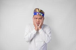 Confused Balinese man in traditional headdress called udeng thinking hard while touching chin with serious expression, isolated by white background photo