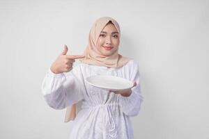 sonriente joven asiático musulmán mujer en un velo hijab señalando a un vacío plato con Copiar espacio terminado él, presentación comida menú para iftar. Ramadán concepto foto