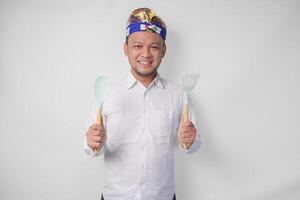 Smiling young Balinese man in white shirt and traditional headdress holding spatula and kitchen cooking utensils photo