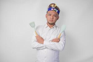 Young Balinese man in white shirt and traditional headdress holding spatula and kitchen cooking utensils with serious expression photo