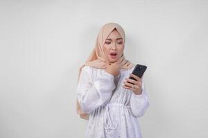 Young Asian Muslim woman wearing white dress and hijab showing shocked expression on her face while holding smartphone, surprised after reading news or gossip, isolated by white background photo